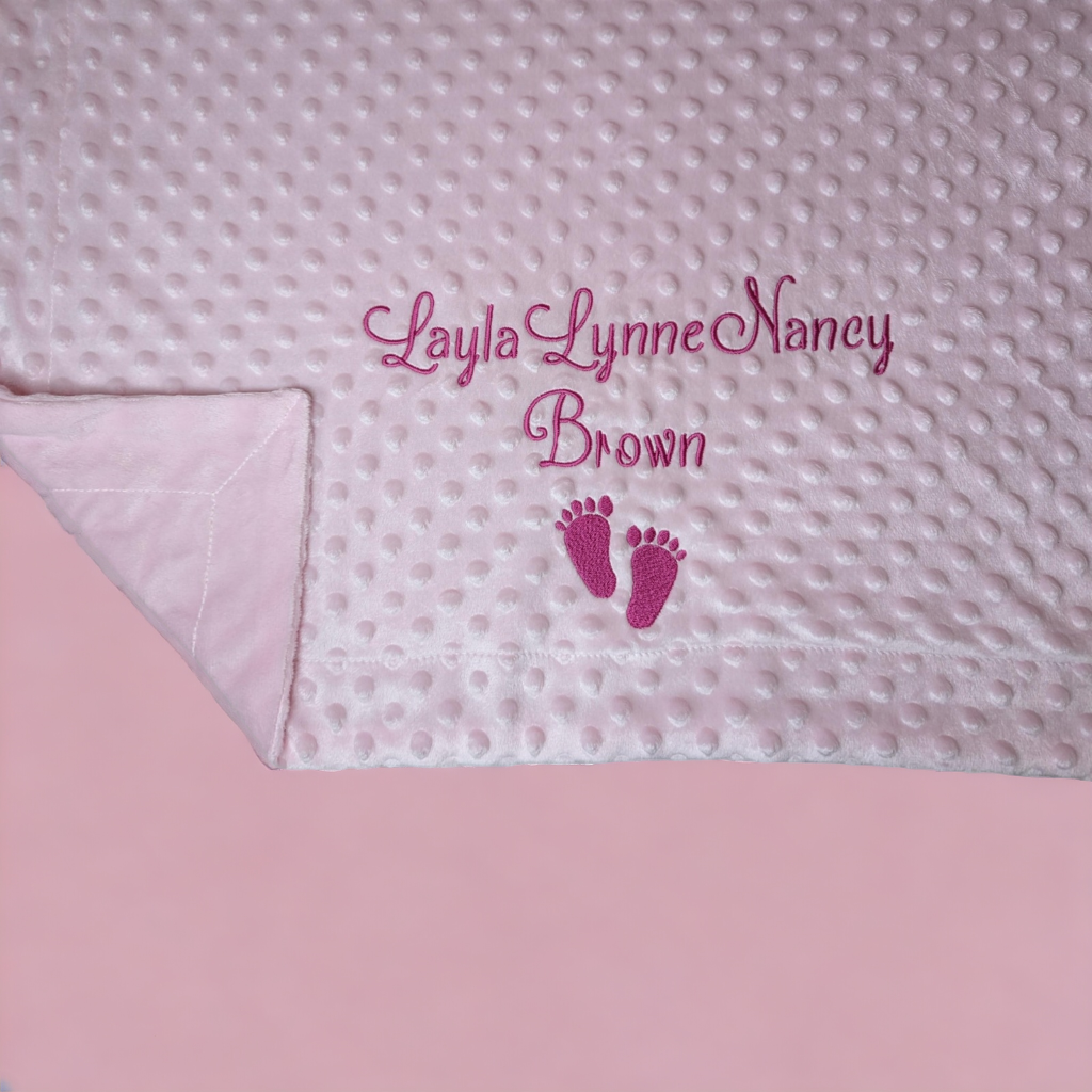 A pink blanket is depicted in this image, featuring a baby's foot and the name "lacy lynne nancy brown. " embroidered on the blanket are the names of the baby.