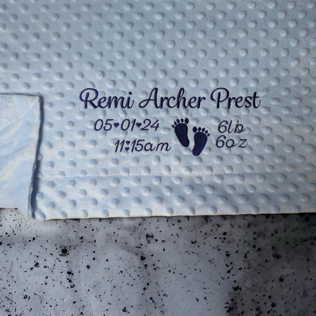 A blue blanket is depicted in this image, featuring a baby's foot and the name "remi archer prest" written on it. The blanket is placed on a grey surface.