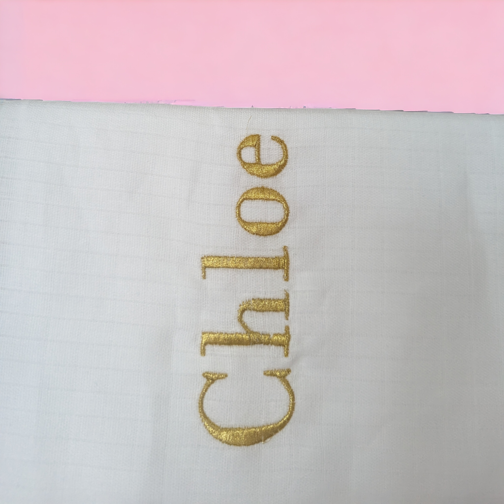 A white muslin is depicted in this image, featuring gold-colored letters and a pink background. The image is slightly blurry in the background.