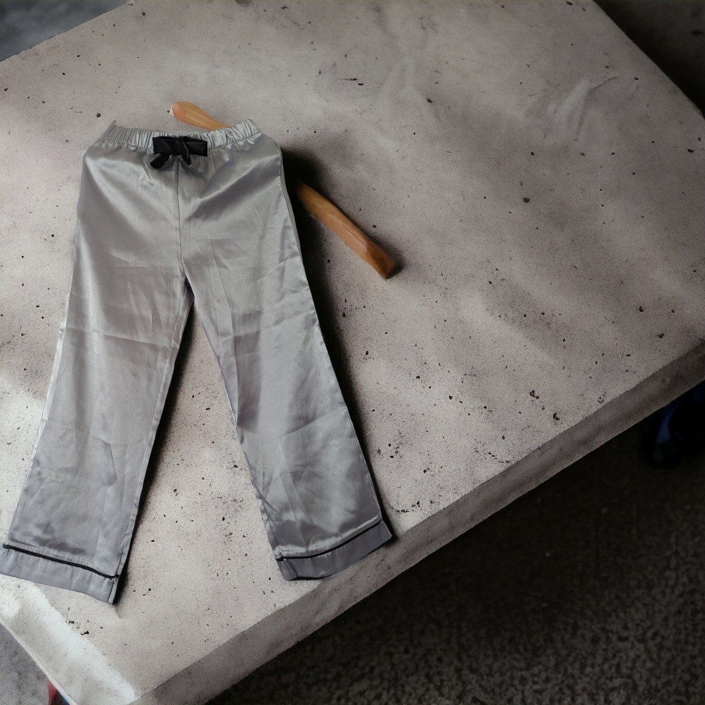 A pair of grey satin pants is depicted in this image, with a wooden stick lying on the floor nearby. The background features a white cloth.