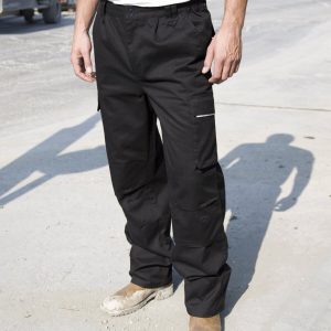 A man is standing in the image, dressed in a white t-shirt and black trousers. The background features a road, a vehicle, and a few unidentified objects.