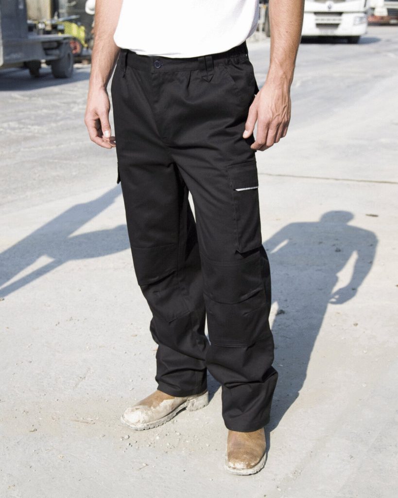 A man is standing in the image, dressed in a white t-shirt and black trousers. The background features a road, a vehicle, and a few unidentified objects.