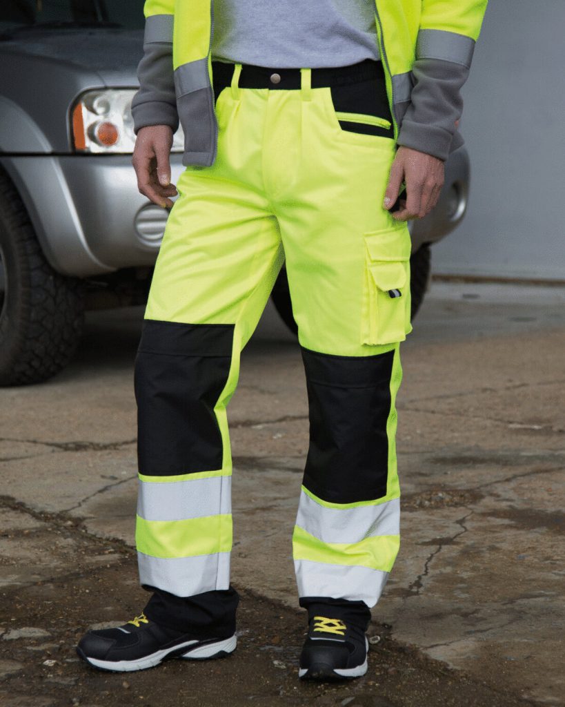 A person is standing in the image, dressed in yellow and black safety cargo trousers. The background features a grey car, a wall, and a few other unidentified objects.