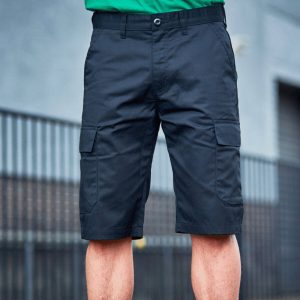 A man is standing in the image, dressed in a green t-shirt and blue shorts. He is holding a mobile phone in his hand. The background features a wall, a gate, and a few plants.