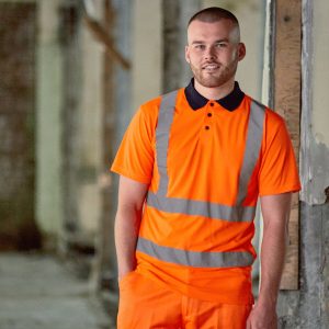 A man is standing in the image, dressed in an orange T-shirt and orange pants. He is smiling and posing for the camera. The background features a wall, pillars, and a wooden object.