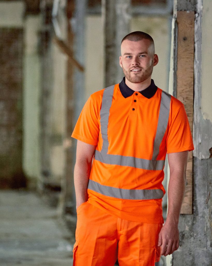 A man is standing in the image, dressed in an orange t-shirt and orange pants. He is smiling and posing for the camera. The background features a wall, pillars, and a wooden object.