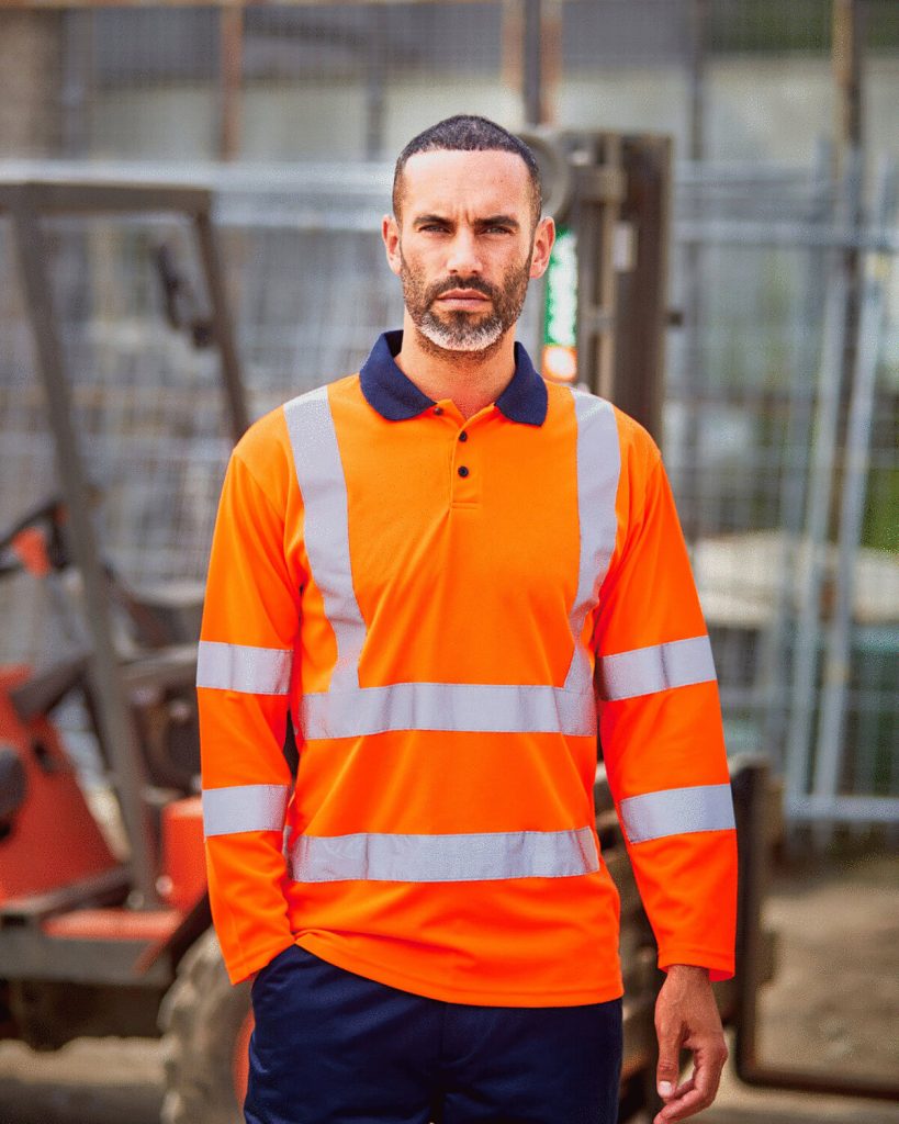 A man is standing in the image, dressed in an orange shirt and blue pants. The background features a forklift, a fence, and a building.