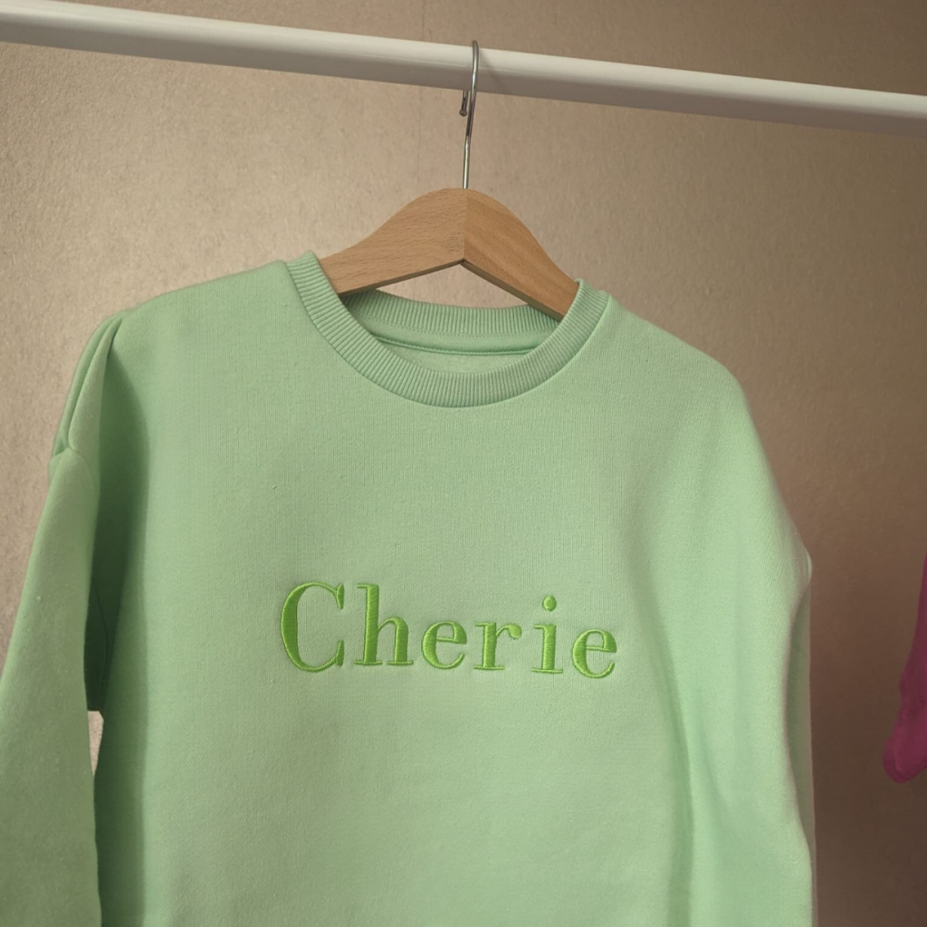 A sweatshirt is depicted in this image, featuring the name "cherie" embroidered on it. The sweatshirt is hanging on a white rod. The background is a cream-colored wall.