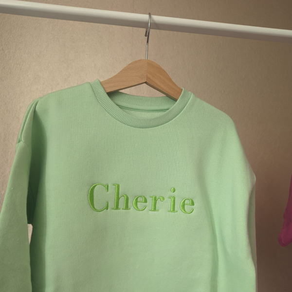 A sweatshirt is depicted in this image, featuring the name "Cherie" embroidered on it. The sweatshirt is hanging on a white rod. The background is a cream-colored wall.