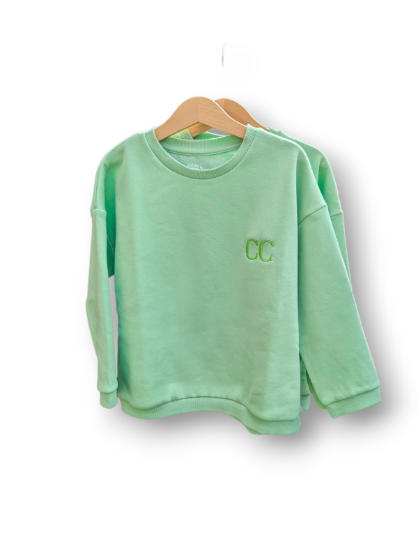 A child's sweatshirt is depicted in this image, featuring a green color and the letters "CC" embroidered on it. The sweatshirt is hanging on a hanger, and a white background is visible.
