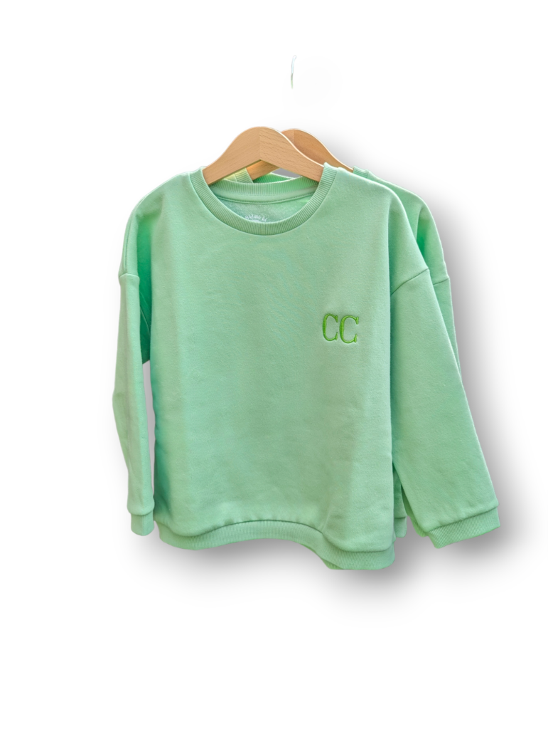 A child's sweatshirt is depicted in this image, featuring a green color and the letters "cc" embroidered on it. The sweatshirt is hanging on a hanger, and a white background is visible.