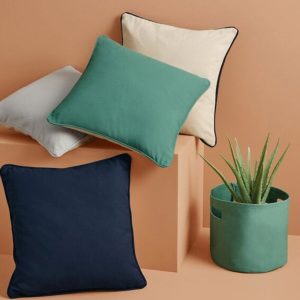 A collection of pillows is arranged on a platform, with a plant situated in a pot nearby. The background features a wall.