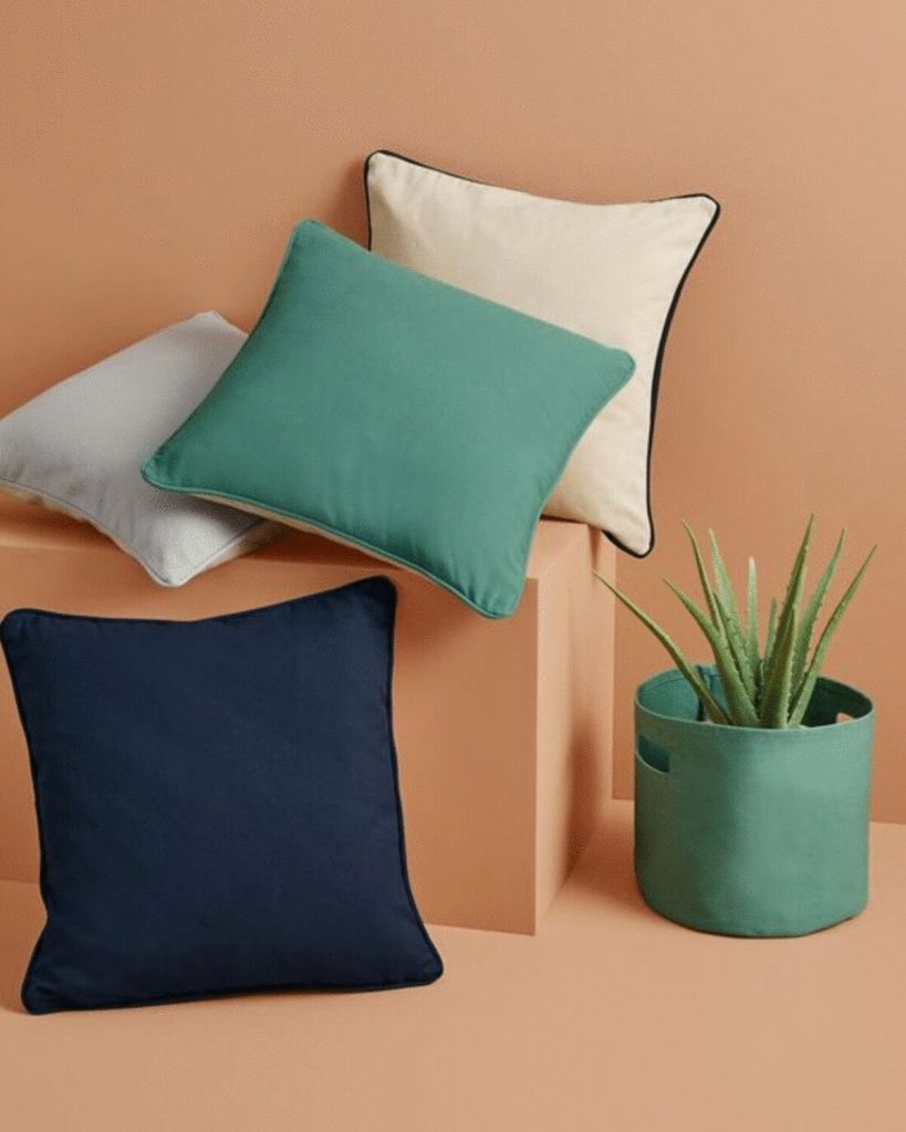 A collection of pillows is arranged on a platform, with a plant situated in a pot nearby. The background features a wall.