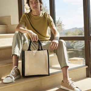 A man is seated on a set of stairs, holding a bag in his hand. The background features a glass window, through which trees and the sky can be seen.