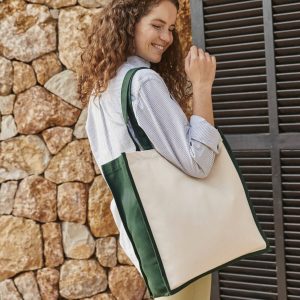 A woman is depicted in this image, holding a green and white canvas tote bag in her hand. She is smiling and appears to be posing for the camera. The background features a stone wall.