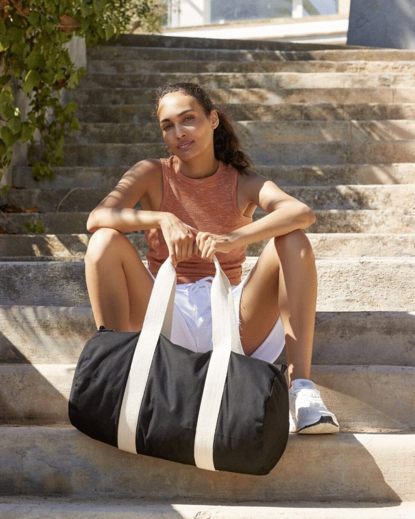 A woman is seated on a set of stairs, holding a black bag in her hand. The background features a wall, plants, and a staircase. The image appears to have been taken during the daytime.