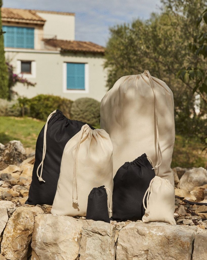 A collection of bags in various colors, including black, white, and cream, are arranged on a rock. The bags are made of cotton and are tied with strings. The background features a building with a blue window and a few trees.