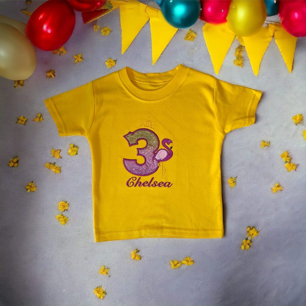 A yellow t-shirt is displayed in the image, featuring a design of a flamingo and the number three. The shirt is placed on a purple and white surface, with yellow balloons and a yellow cloth in the background.