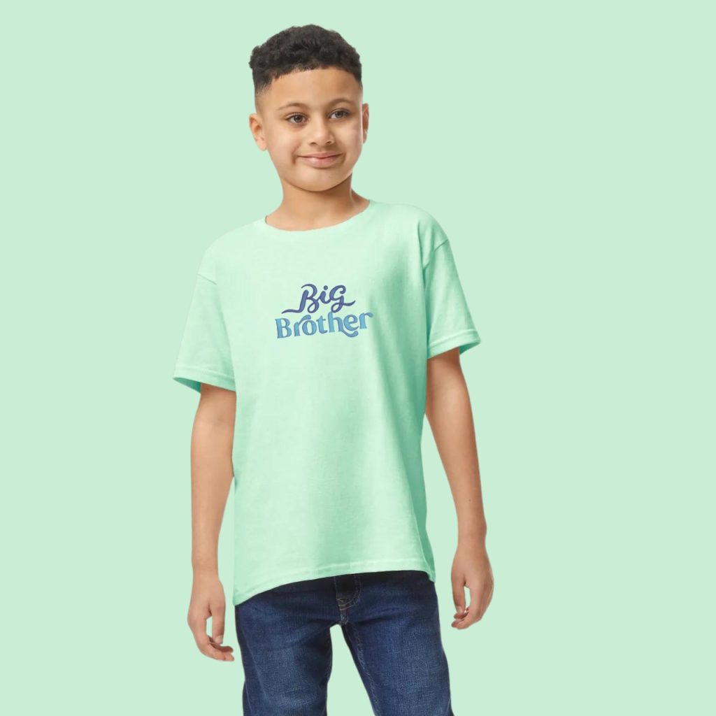 A boy is standing in the image, dressed in a green t-shirt with the words "big brother" written on it. The background is white.