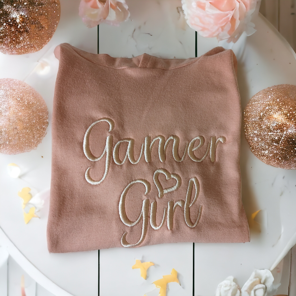 A pink-colored hoodie is depicted in this image, with the word "gamer girl" written on it. The hoodie is placed on a white surface, which also holds a few decorative items.