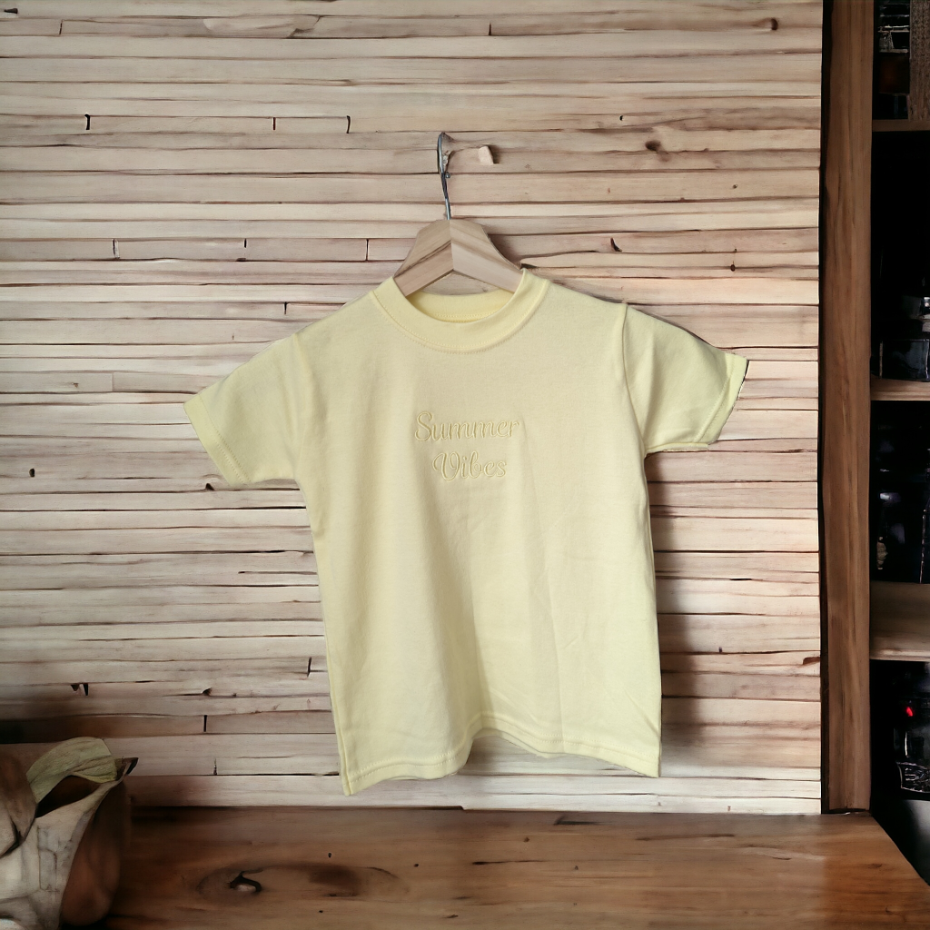 A yellow t-shirt is displayed in the image, featuring the word "summer" written in white letters. The shirt is hanging on a hanger, and a wooden wall is visible in the background.