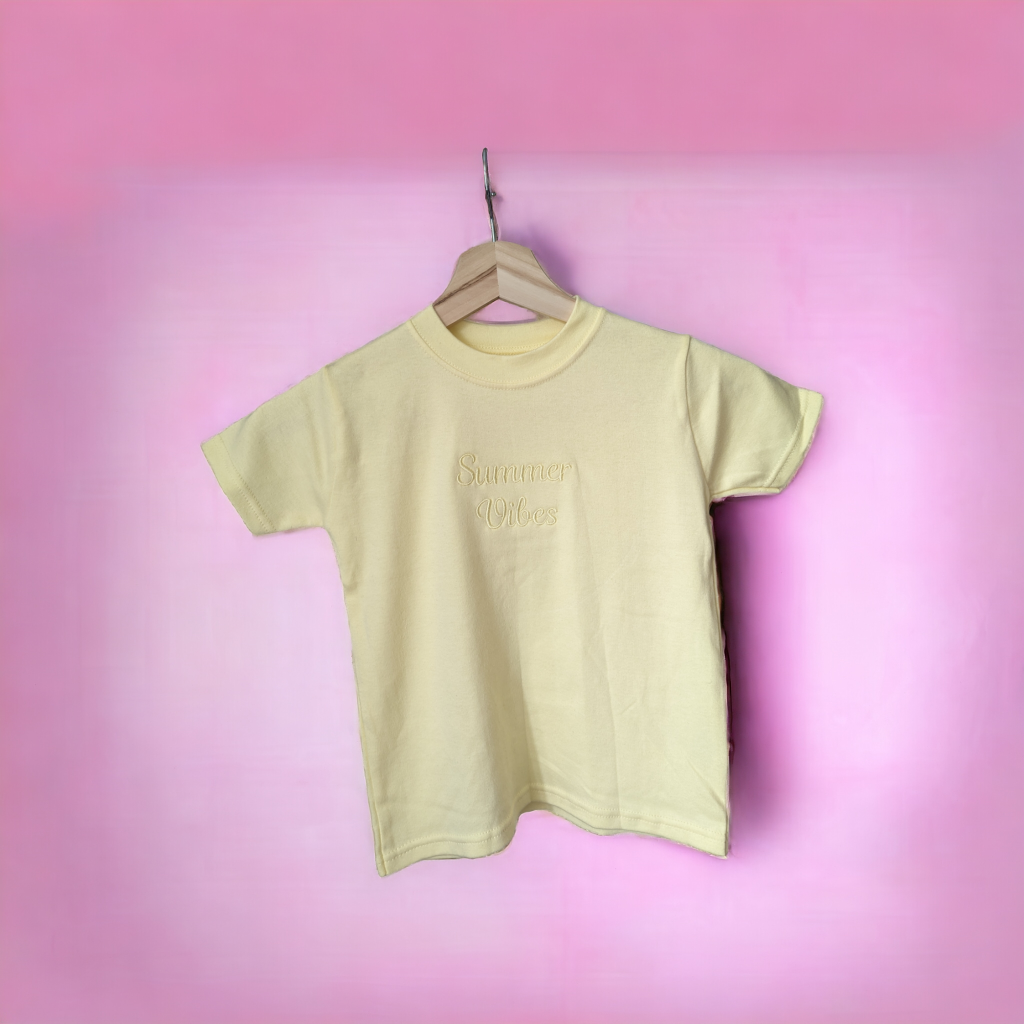 A yellow t-shirt is displayed in the image, featuring the word "lemon" written on it. The shirt is hanging on a hanger, and a pink background is visible behind it.