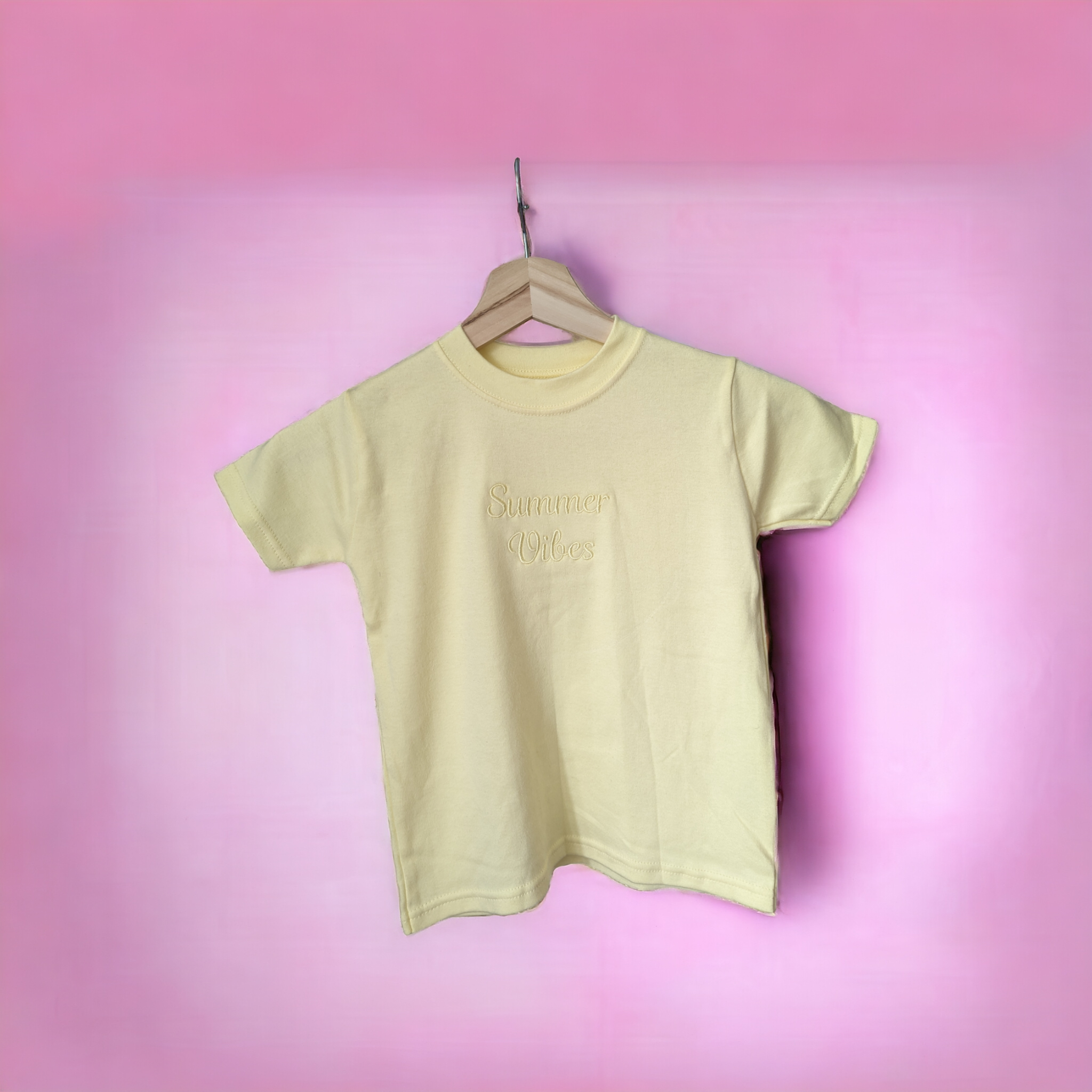 A yellow t-shirt is displayed in the image, featuring the word "Lemon" written on it. The shirt is hanging on a hanger, and a pink background is visible behind it.