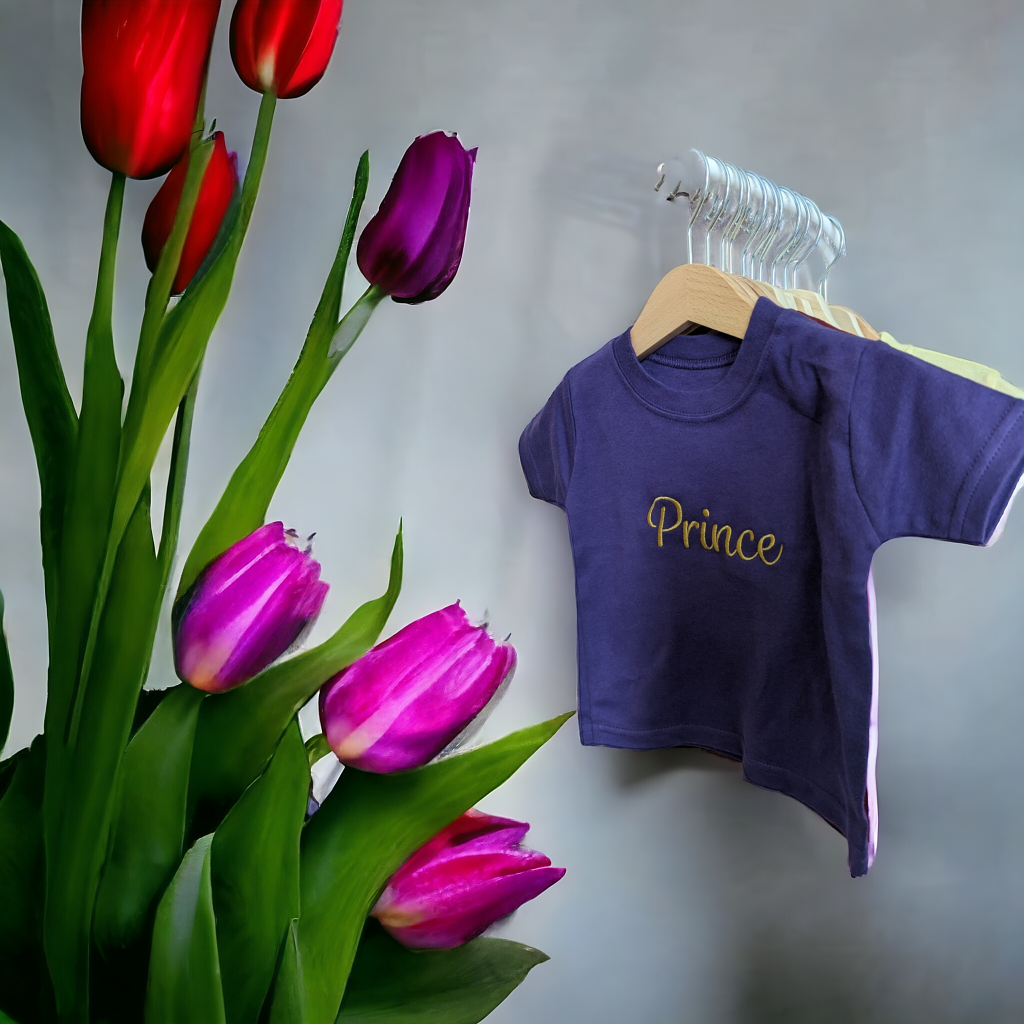 A shirt is displayed in the image, featuring the name "prince. " the shirt is hanging on a hanger, and a flower vase is situated nearby. The background is composed of a wall.