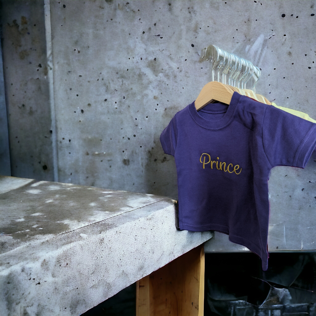 A purple t-shirt is displayed in the image, hanging on a hanger. The shirt features the name "prince" written on it. The background is a grey wall.