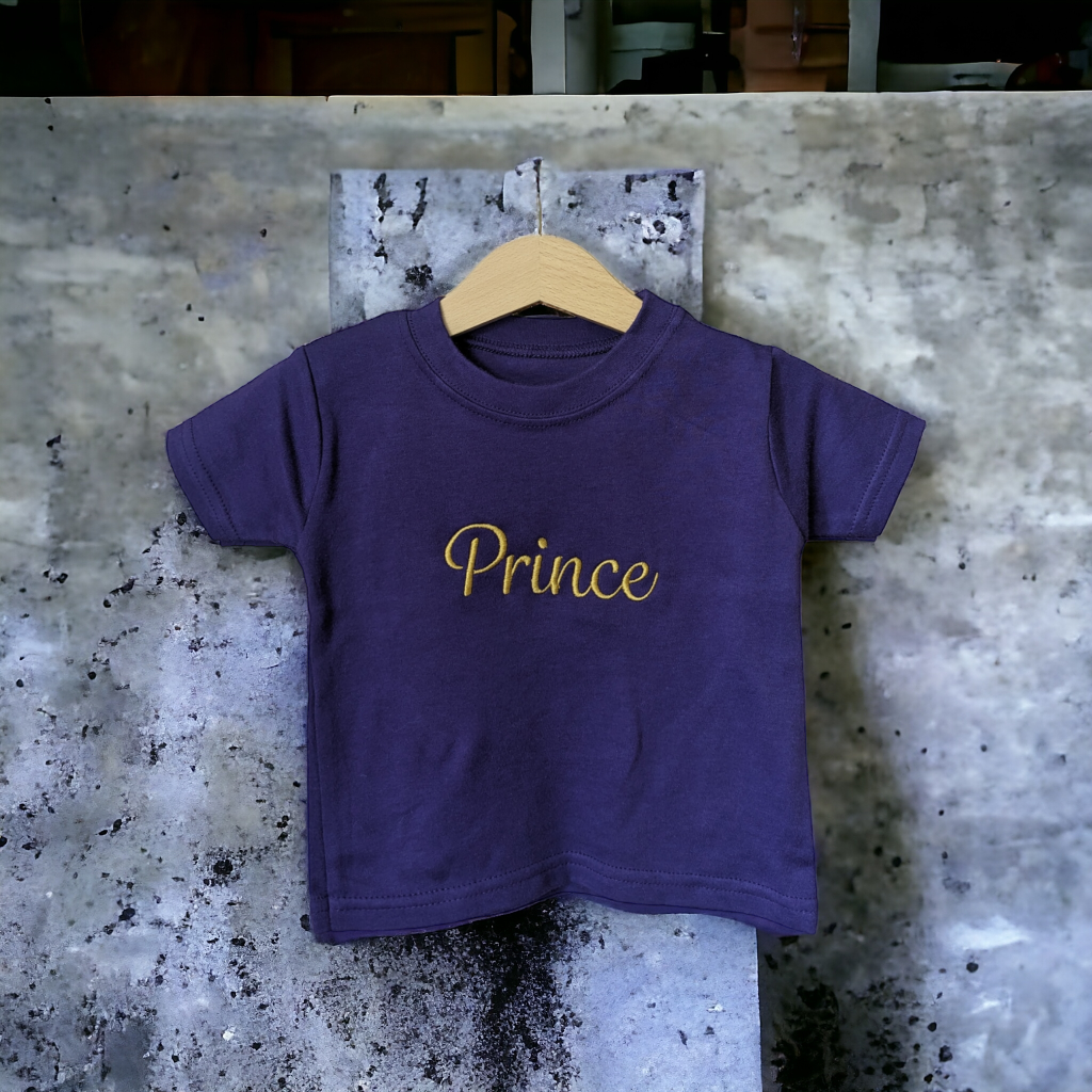 A child's purple t-shirt is displayed in the image, featuring the name "prince" written in yellow letters. The shirt is hanging on a wooden hanger, which is attached to a wall.