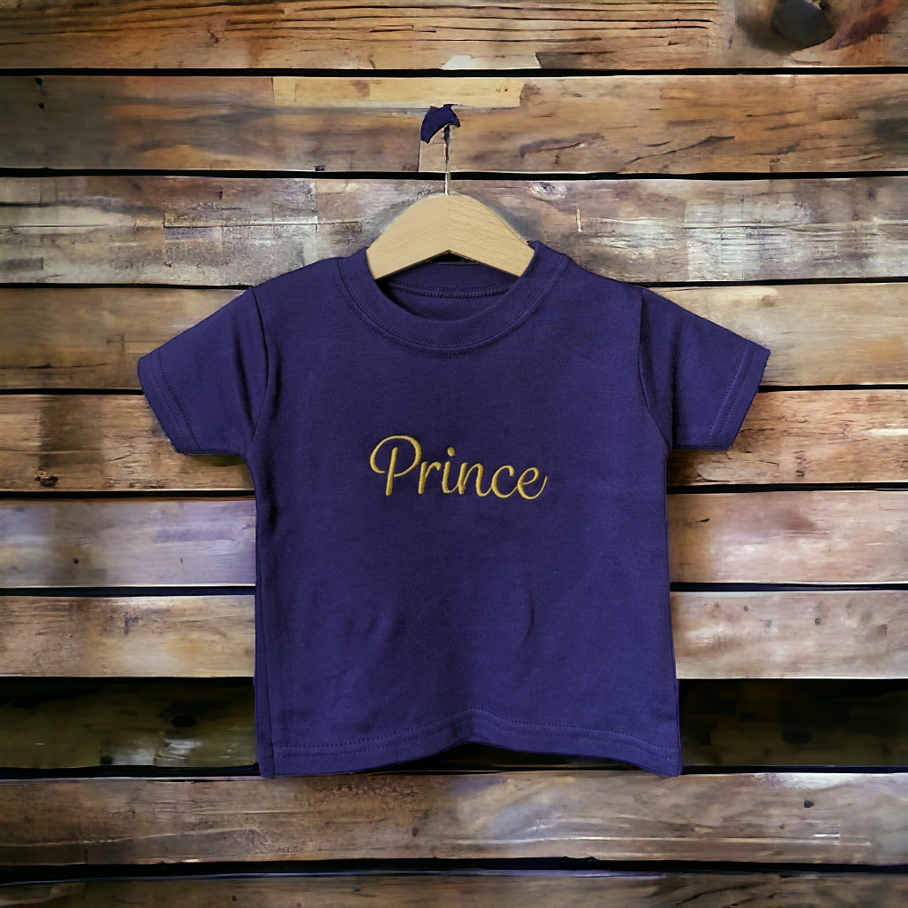 A purple t-shirt is displayed in the image, featuring the name "prince" written in yellow letters. The shirt is hanging on a wooden hanger, which is placed on a wooden surface.