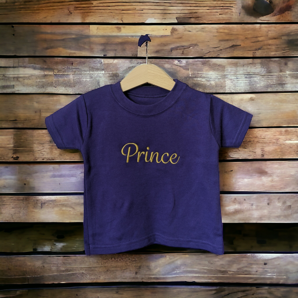A purple t-shirt is displayed in the image, featuring the name "Prince" written in yellow letters. The shirt is hanging on a wooden hanger, which is placed on a wooden surface.