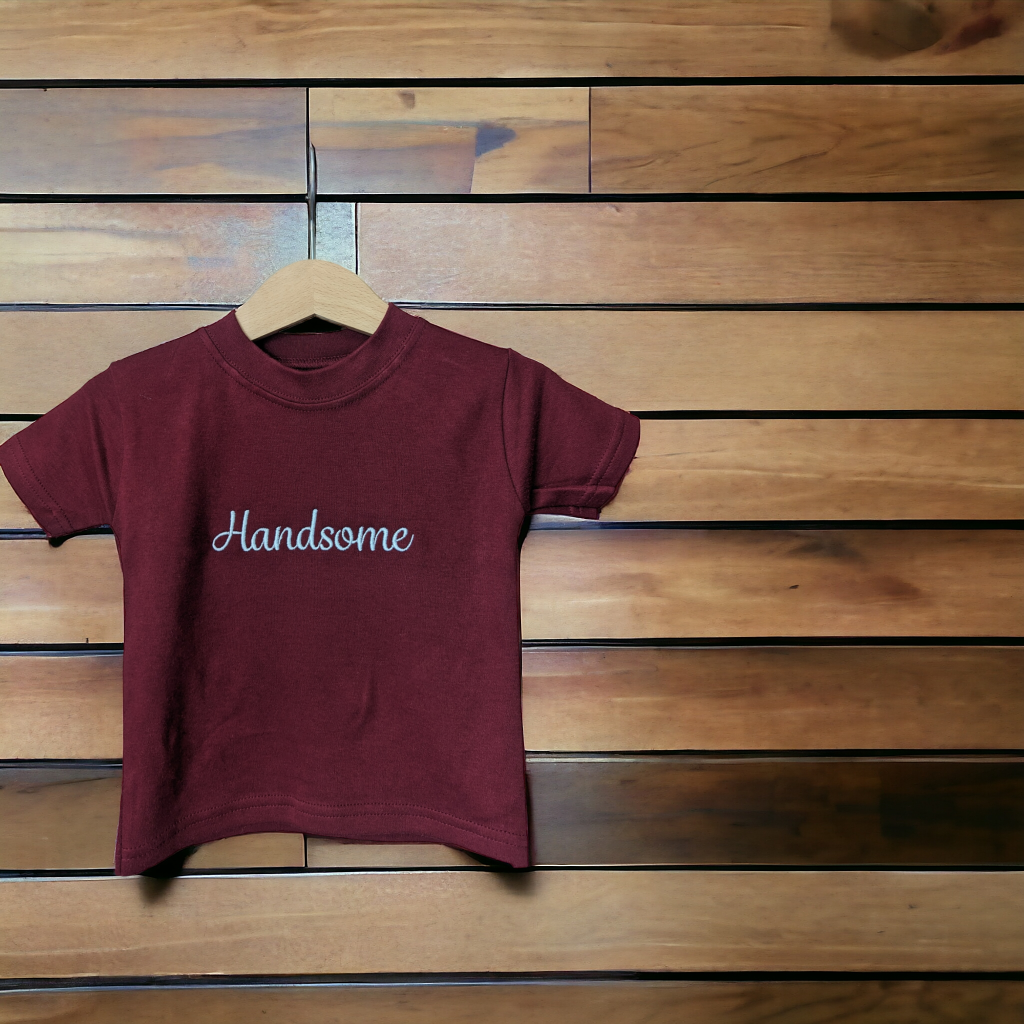 A maroon-colored t-shirt is displayed in the image, featuring the word "handsome" written in white letters. The shirt is hanging on a hanger, attached to a wooden wall.