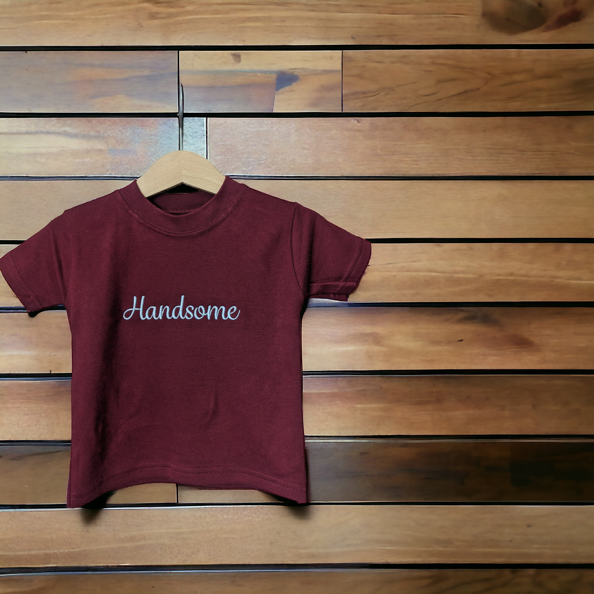 A maroon-colored t-shirt is displayed in the image, featuring the word "handsome" written in white letters. The shirt is hanging on a hanger, attached to a wooden wall.