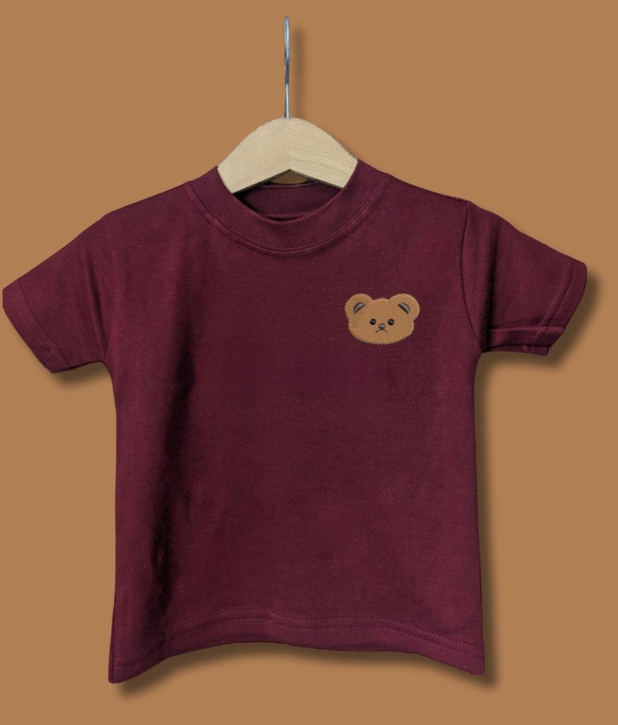 A maroon-colored t-shirt is displayed in the image, featuring a teddy bear's face. The teddy bear is situated in the center of the shirt, and a wooden hanger is visible behind it.