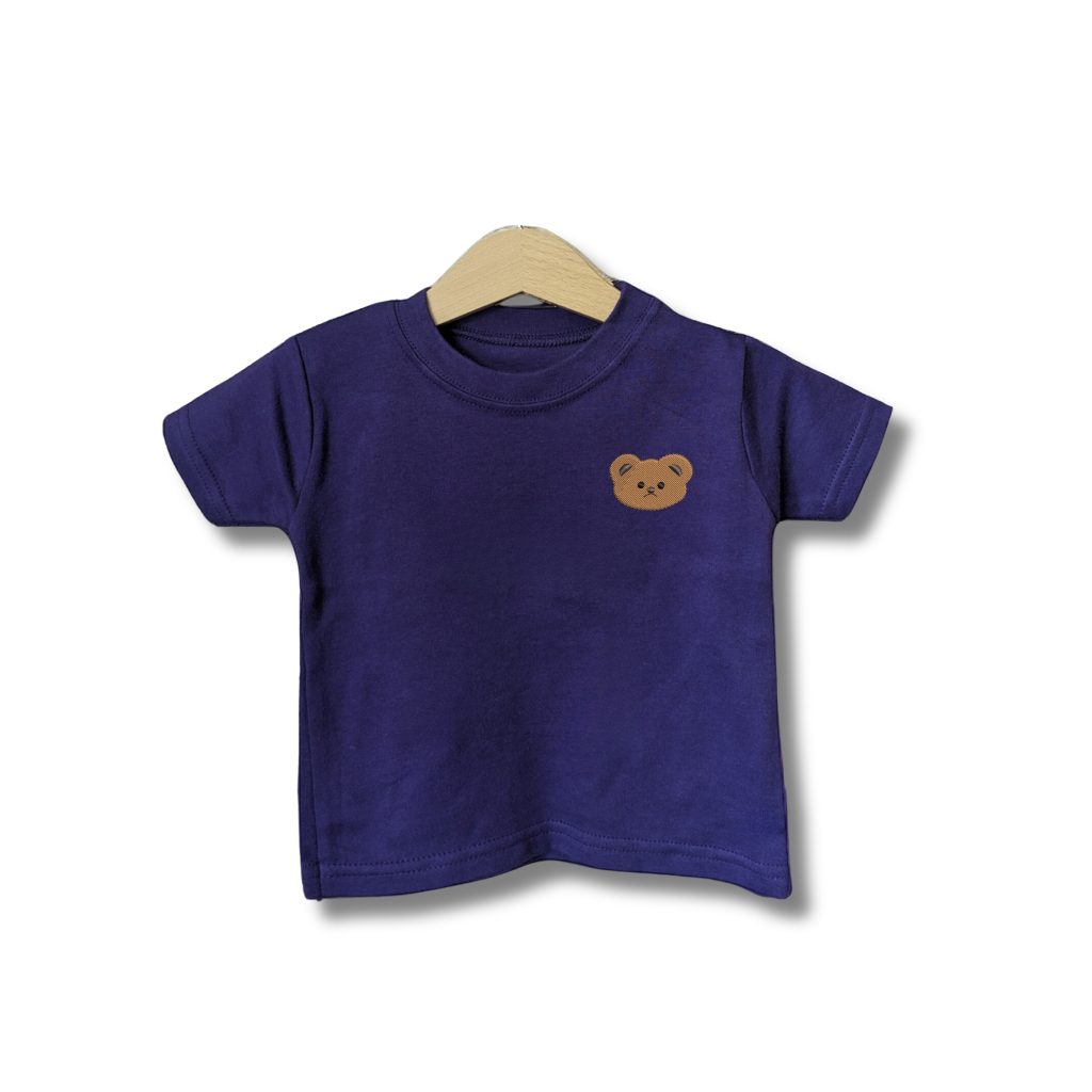 A baby is depicted in this image, wearing a purple t-shirt with a brown bear face on it. The shirt is hanging on a wooden hanger, and the background is white.