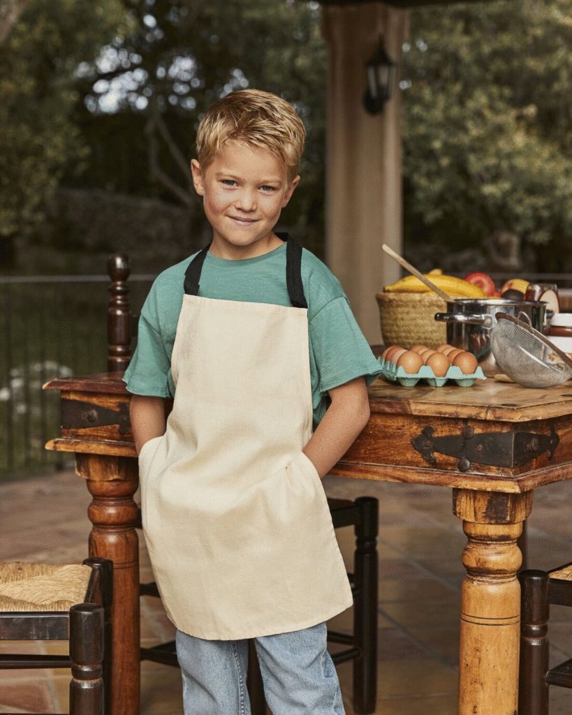 A child is standing in front of a table, which is adorned with a basket of fruits, a bowl, a pan, and other items. The child is wearing a white apron with black bands. The background features a railing, trees, and the sky.