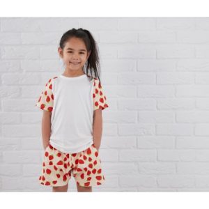 A girl is standing in the image, dressed in a white shirt and shorts with a red and green strawberry pattern. The background is a white brick wall.