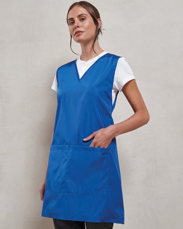 A woman is standing in the image, dressed in a blue tunic, with a white t-shirt and black pants. The background is a white wall.