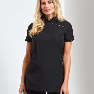 A woman is standing in the image, dressed in a black tunic. She is holding a book and a bag in her hands. The background is white, and a wall is visible.