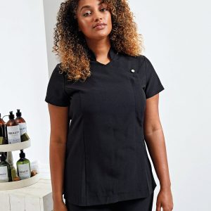 A woman is standing in the image, dressed in a black tunic. In front of her, a table is arranged with bottles. The background is composed of a wall.