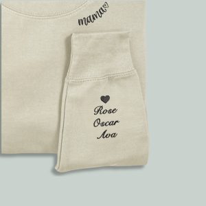 A white sweatshirt is displayed in the image, featuring a heart and names embroidered on the sleeves. The sweatshirt is adorned with a black heart on the left side and a name on the right side.