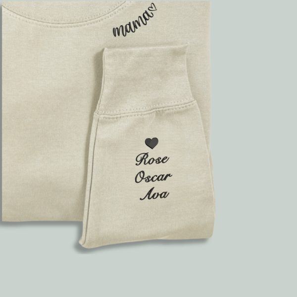 A white sweatshirt is displayed in the image, featuring a heart and names embroidered on the sleeves. The sweatshirt is adorned with a black heart on the left side and a name on the right side.